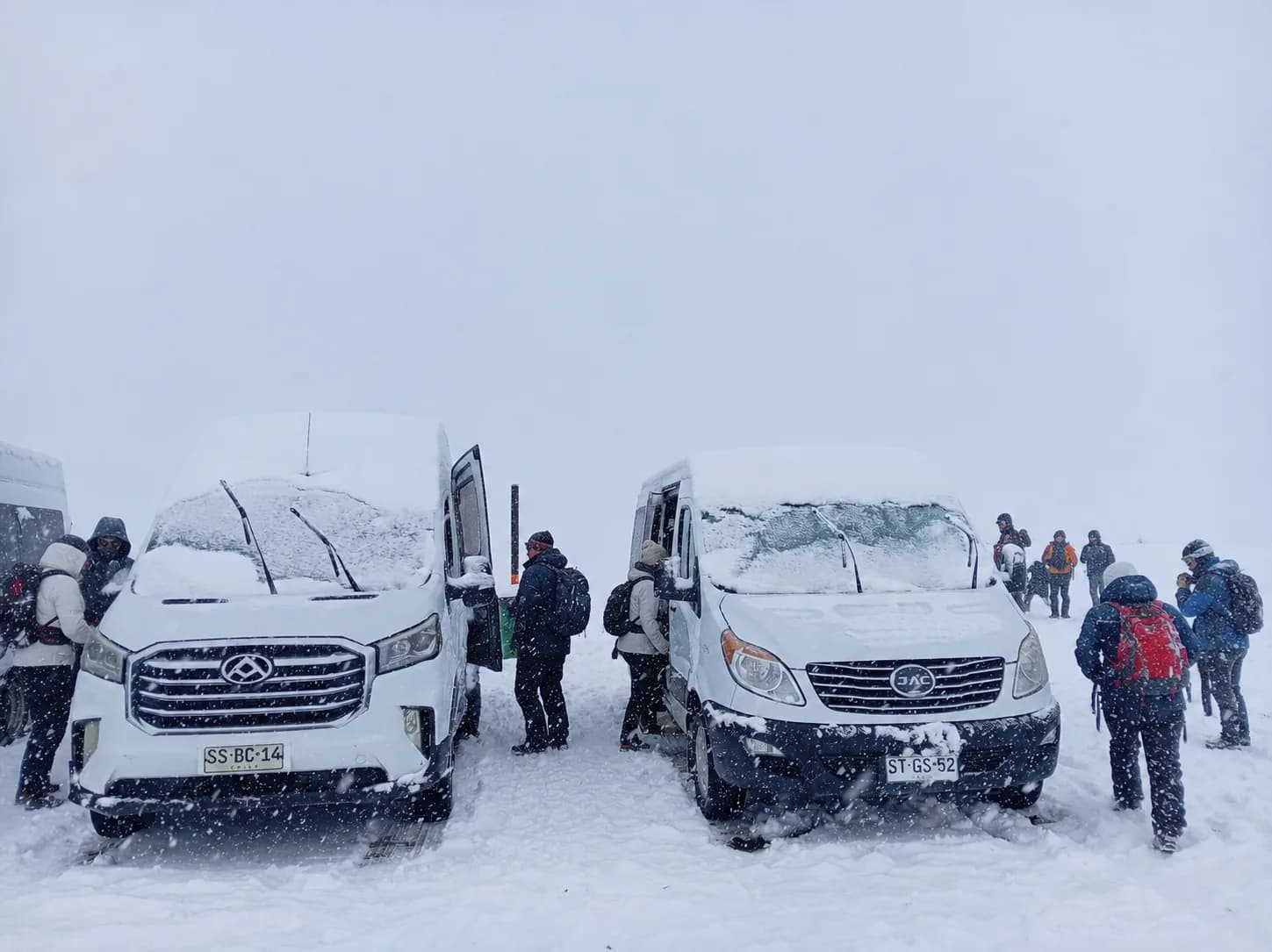 Dos vans con nieve en el techo y un grupo de personas con ropa de abrigo y mochilas durante una nevada